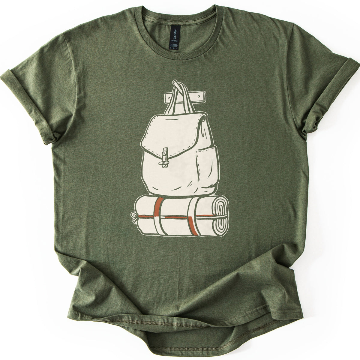 Backpack And Bedroll Comfort Colors T-shirt in Military Green, laid flat. A cream illustration of a canvas backpack with a toggle-clasp flap hangs by its handles from a wall peg above a rolled bedroll tied with rust-red straps. No words. Cuffed sleeves on a white surface.