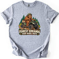 Forest Errands Four Wheel Drive Comfort Colors T-shirt in Sport Grey, laid flat. A grinning Bigfoot in a green helmet rides a green four-wheeler through mud past pine trees, a sack of pinecones on the back, above the words Forest Errands Four Wheel Drive. Shown on a white background.