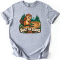 Built For Berms Comfort Colors T-shirt in Sport Grey, laid flat. A smiling cartoon beaver rakes a curving dirt berm with pine trees and a snowy mountain behind, above a wooden sign reading Built For Berms MTB. The sleeves are cuffed on a white background.