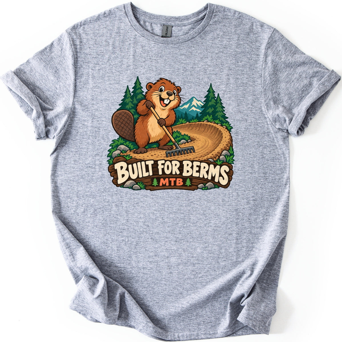 Built For Berms Comfort Colors T-shirt in Sport Grey, laid flat. A smiling cartoon beaver rakes a curving dirt berm with pine trees and a snowy mountain behind, above a wooden sign reading Built For Berms MTB. The sleeves are cuffed on a white background.