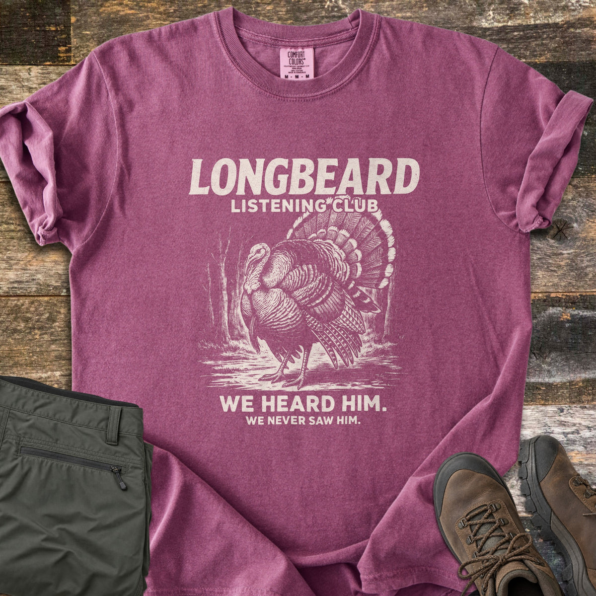 Longbeard Listening Club Comfort Colors T-shirt in Berry, laid flat. A cream engraving of a strutting wild turkey in the woods sits under Longbeard Listening Club, with We heard him. We never saw him. below. Olive hiking pants and brown boots lie beside it on weathered wood.