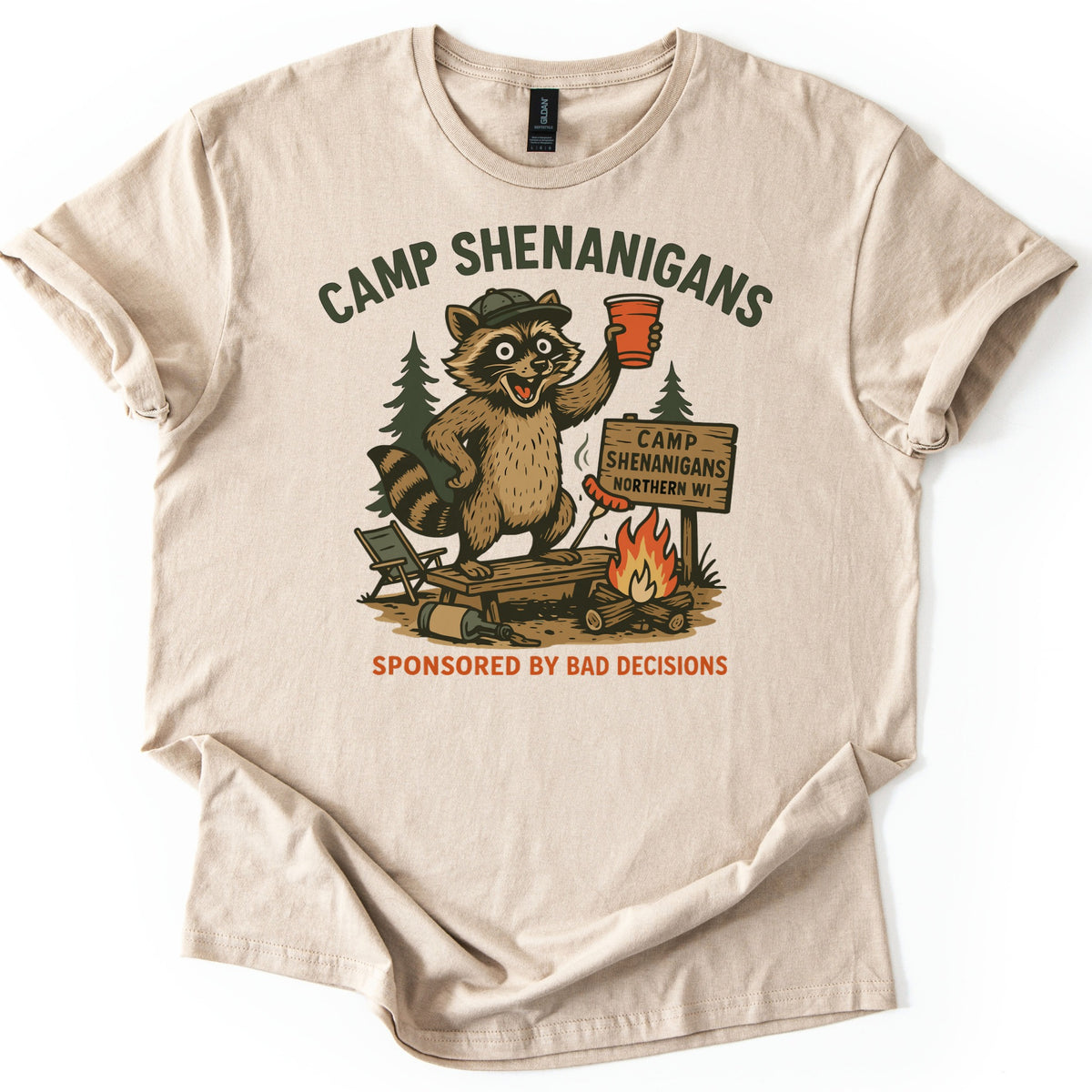 Camp Shenanigans Comfort Colors T-shirt in Sand, laid flat. A raccoon in a cap raises a red cup on a bench by a campfire and a sign reading Camp Shenanigans Northern WI, below Camp Shenanigans and above Sponsored by Bad Decisions. Shown on a white background.