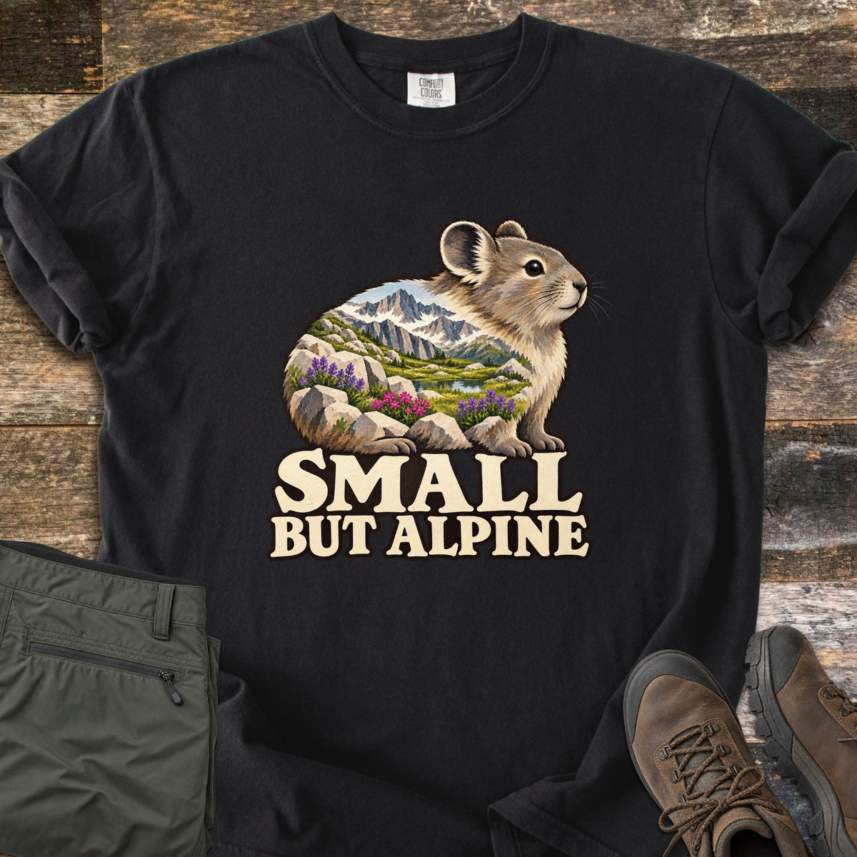 Small But Alpine Comfort Colors T-shirt in Black, laid flat. A pika whose body is filled with a mountain scene of snowy peaks, a lake, and purple and pink wildflowers among rocks sits above the words Small But Alpine. Green hiking pants and brown boots rest on weathered wood.
