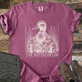 The Waterfowler Comfort Colors T-shirt in Berry, laid flat. A cream engraving-style mallard in a camo jacket with a duck call on a cord stands in marsh grass above the words The Waterfowler. Folded olive pants and brown hiking boots sit beside it on weathered wood.