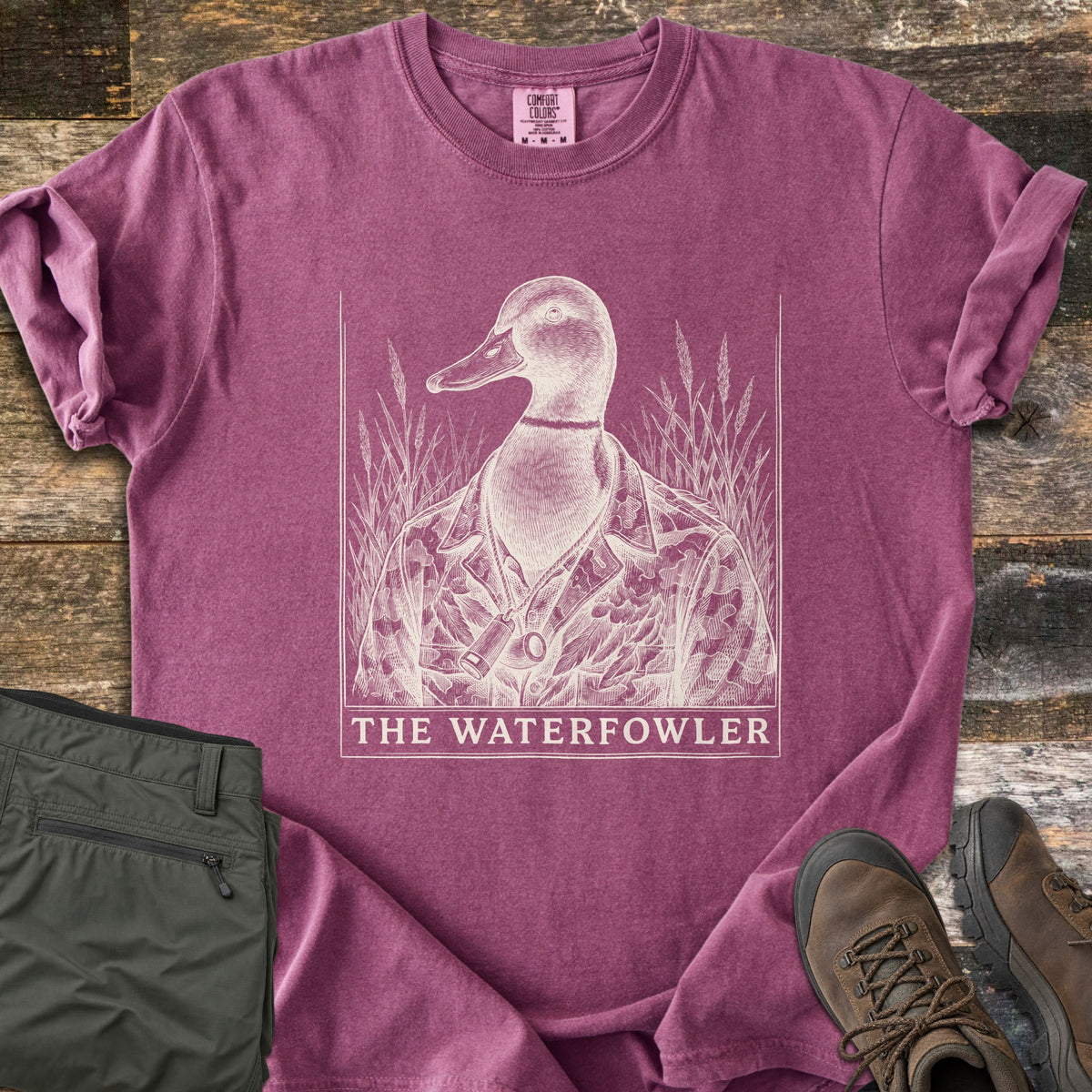The Waterfowler Comfort Colors T-shirt in Berry, laid flat. A cream engraving-style mallard in a camo jacket with a duck call on a cord stands in marsh grass above the words The Waterfowler. Folded olive pants and brown hiking boots sit beside it on weathered wood.