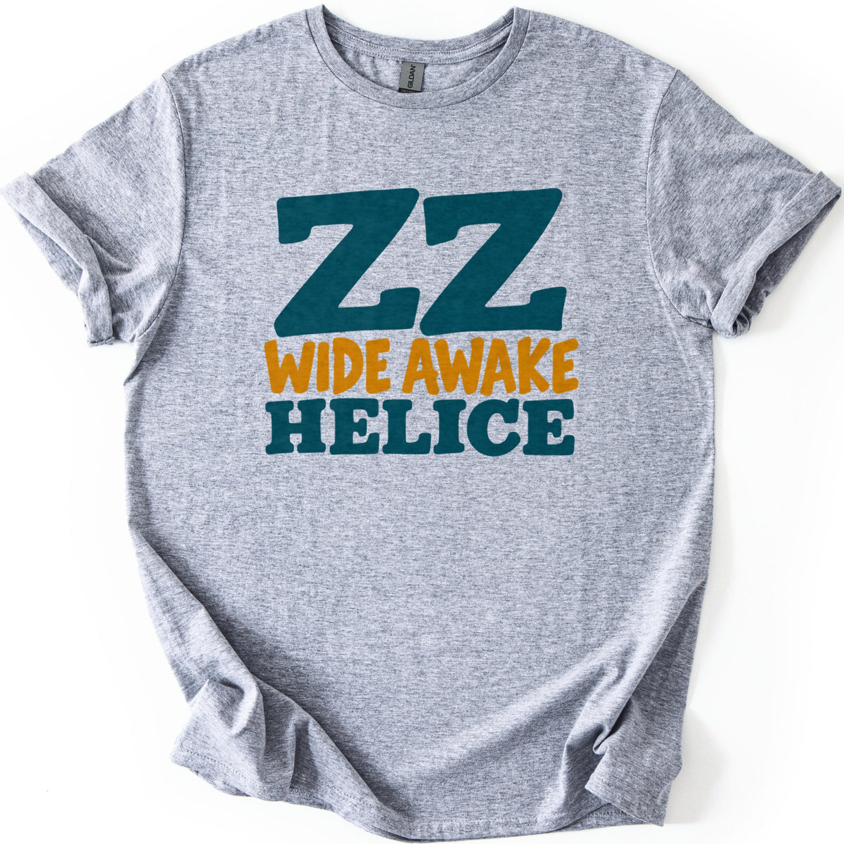 ZZ Wide Awake Comfort Colors T-shirt in Sport Grey, laid flat. Large dark teal letters ZZ sit above WIDE AWAKE in mustard yellow and HELICE in bold dark teal. The shirt lies on a white background with its sleeves cuffed and the hem loosely bunched.