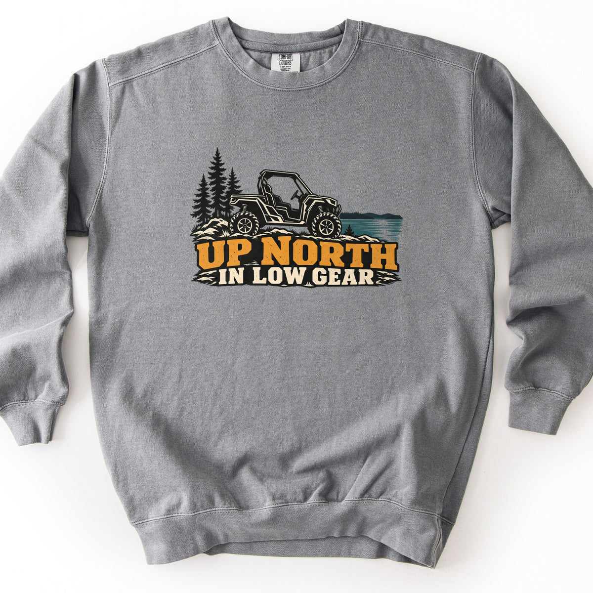 Up North in Low Gear Comfort Colors crewneck sweatshirt in Grey, laid flat. A black and cream side-by-side off-road vehicle sits on rocks between pine trees and a teal lake above the words UP NORTH IN LOW GEAR in gold and cream. It lies on a white background.