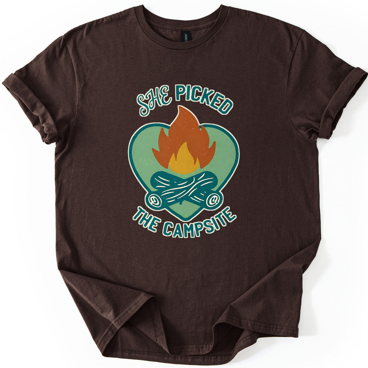 She Picked The Campsite Comfort Colors T-shirt in Dark Chocolate, laid flat. An orange and yellow campfire on crossed teal logs inside a sage green heart, with the words SHE PICKED above and THE CAMPSITE below. The shirt lies on a white surface with its sleeves cuffed.