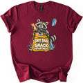 Snack Department Comfort Colors T-shirt in Maroon, laid flat. A smiling raccoon in a blue life vest holds up a snack bar beside a blue paddle, leaning on a yellow dry bag full of snacks printed with Dry Bag Snack Department. The shirt lies on a white background.