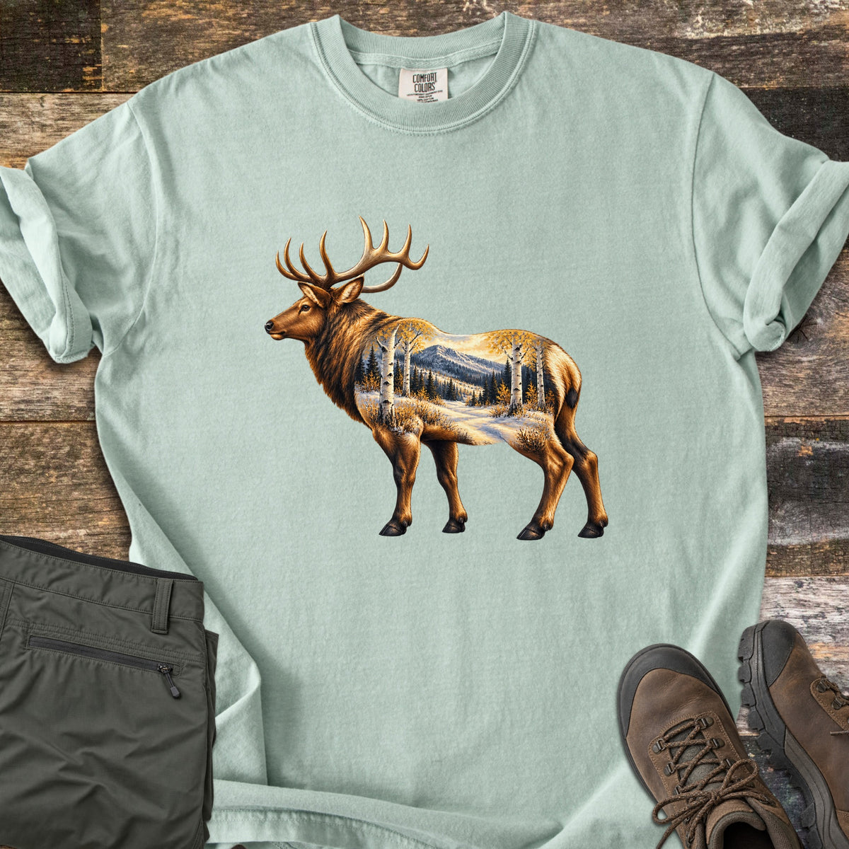 Aspen Pass Elk Comfort Colors T-shirt in Bay, laid flat. A bull elk in profile, its body filled with a snowy mountain scene of aspen trees and pines in golden light. Sleeves rolled, on weathered wood with folded olive hiking pants and brown hiking boots.