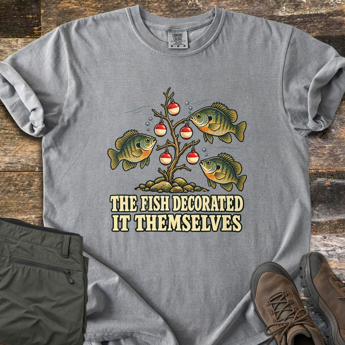 The Fish Decorated It Themselves Comfort Colors T-shirt in Grey, laid flat. Three bluegill fish hang red and white fishing bobbers like ornaments on a bare underwater branch above the words The Fish Decorated It Themselves. Olive pants and brown hiking boots sit on weathered wood.