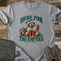 Here for the Empties Comfort Colors T-shirt in Grey, laid flat. A smiling raccoon sits in grass holding red shotgun shells between the words Here for the Empties, with Clay Crew below. Olive cargo pants and brown hiking boots lie beside it on weathered wood.