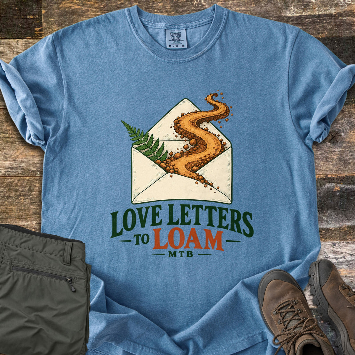 Love Letters to Loam Comfort Colors T-shirt in Blue Jean, laid flat. An open envelope with a fern and a winding dirt trail spilling out, above the words Love Letters to Loam MTB. Shown on weathered wood with folded olive pants and brown hiking boots.