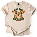 Wide Eyed for Doubles Comfort Colors T-shirt in Sand, laid flat. A cartoon owl with wide yellow eyes glances at two orange clay targets under the arched words Wide Eyed, with For Doubles and Skeet below. The shirt lies on a white surface with sleeves cuffed.
