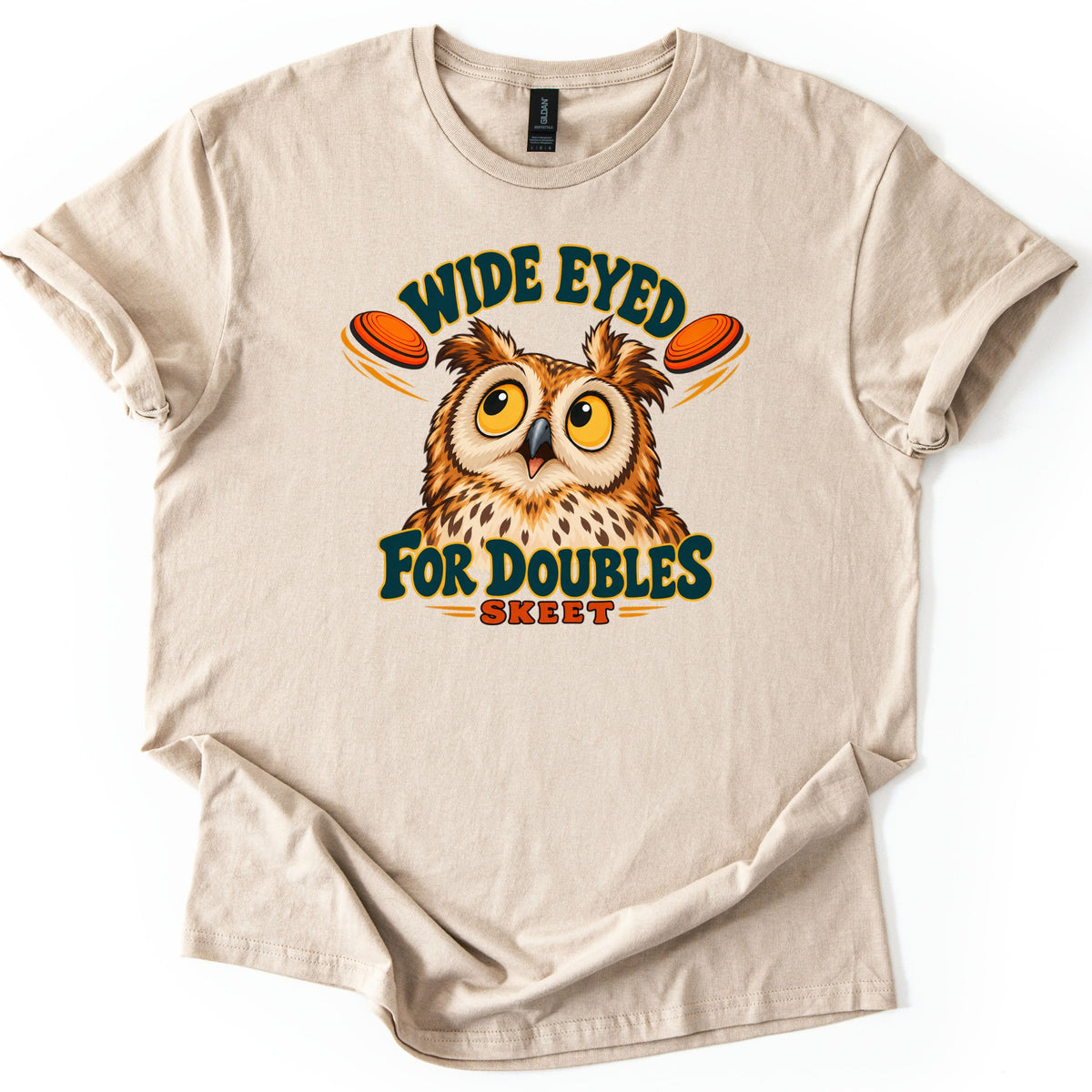 Wide Eyed for Doubles Comfort Colors T-shirt in Sand, laid flat. A cartoon owl with wide yellow eyes glances at two orange clay targets under the arched words Wide Eyed, with For Doubles and Skeet below. The shirt lies on a white surface with sleeves cuffed.