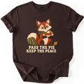 Pass the Pie Keep the Peace Comfort Colors T-shirt in Dark Chocolate, laid flat. A stitched patchwork fox with a green plaid tail and bandana holds a plate of pumpkin pie beside an oak leaf, above the words Pass the Pie Keep the Peace. The sleeves are rolled on a white background.