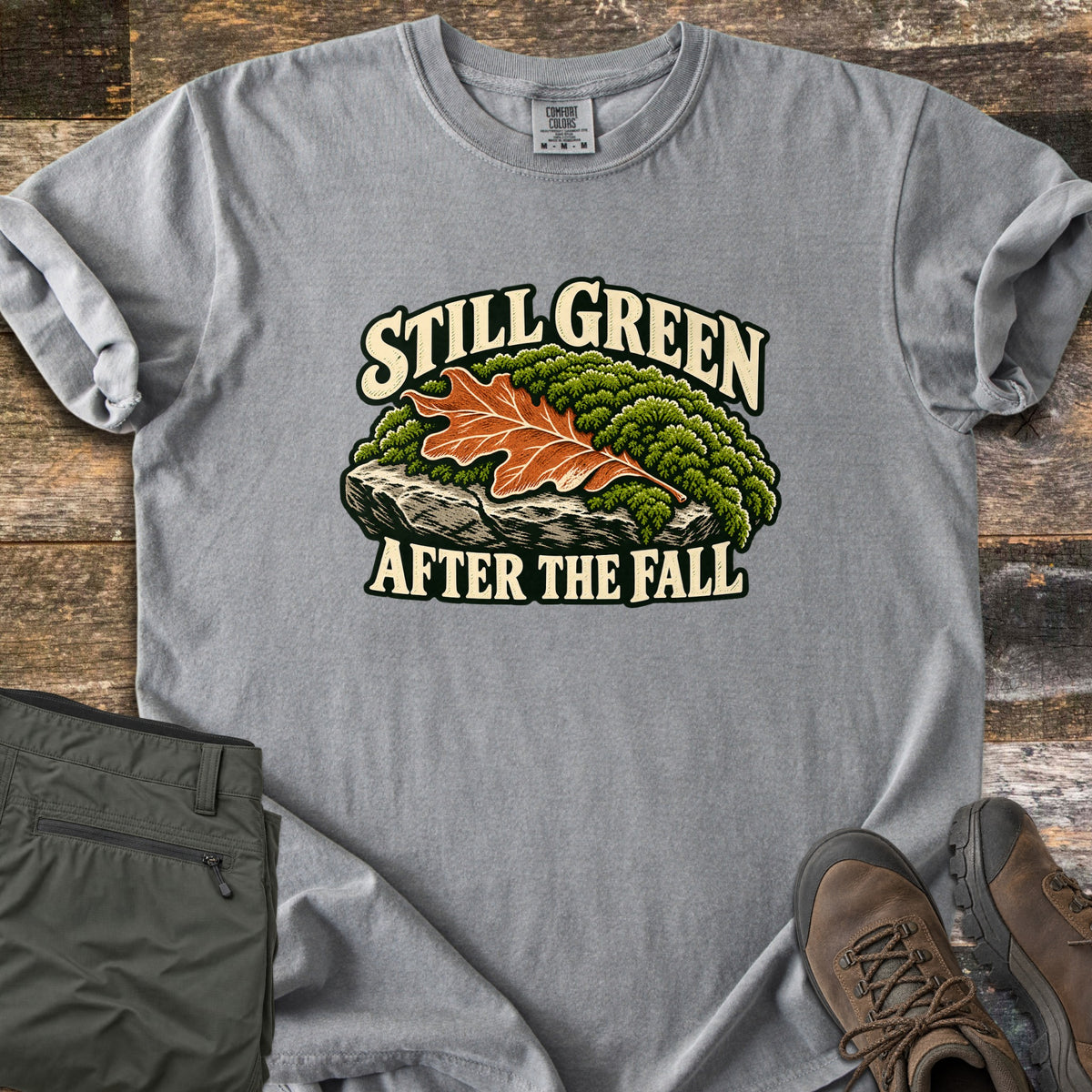 Still Green After the Fall Comfort Colors T-shirt in Grey, laid flat. An orange oak leaf rests on green moss atop a gray rock, with Still Green above and After the Fall below in cream letters. Folded olive hiking pants and brown hiking boots lie beside it on weathered wood planks.