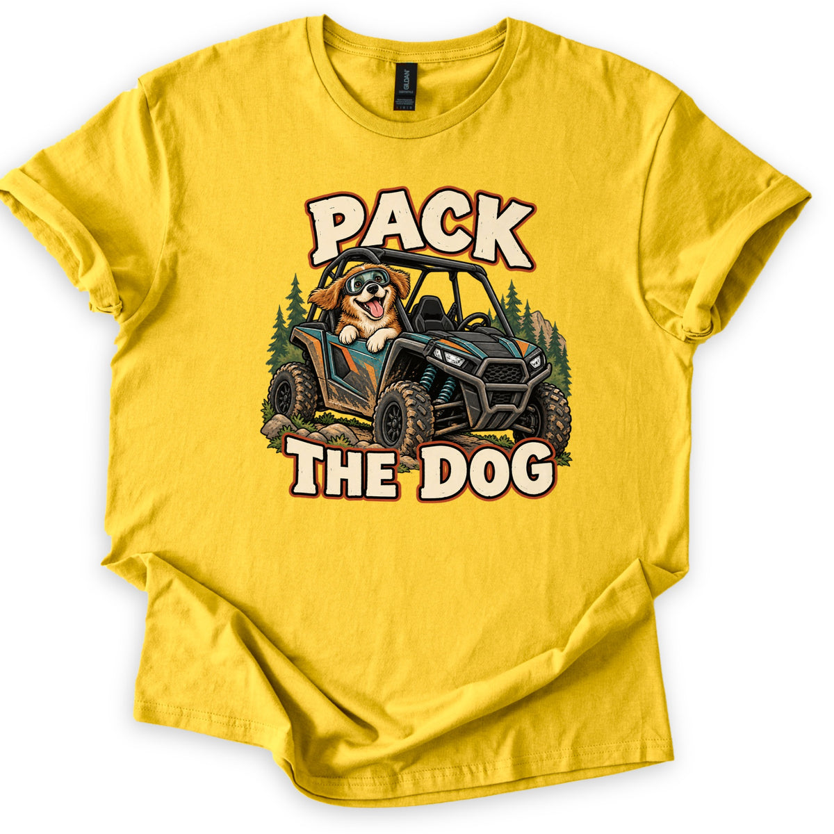 Pack the Dog Comfort Colors T-shirt in Daisy, laid flat. A cartoon dog in goggles rides a muddy teal and black side-by-side UTV past pine trees, between the words PACK and THE DOG in cream letters. The shirt has cuffed sleeves on a white background.