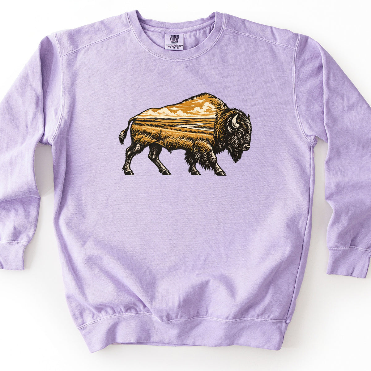 Prairie Bison Comfort Colors crewneck sweatshirt in Orchid, laid flat. A woodcut-style bison walks to the right, its body filled with a golden prairie scene of tall grass, a winding river and clouds. There are no words. The sweatshirt lies on a white surface.