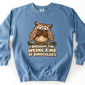 I Brought The Wrong Kind Of Binoculars Comfort Colors crewneck sweatshirt in Blue Jean, laid flat. A cartoon brown owl perched on a mossy branch holds two pine cones up to its eyes like binoculars, above the words I Brought The Wrong Kind Of Binoculars. Shown on a white background.