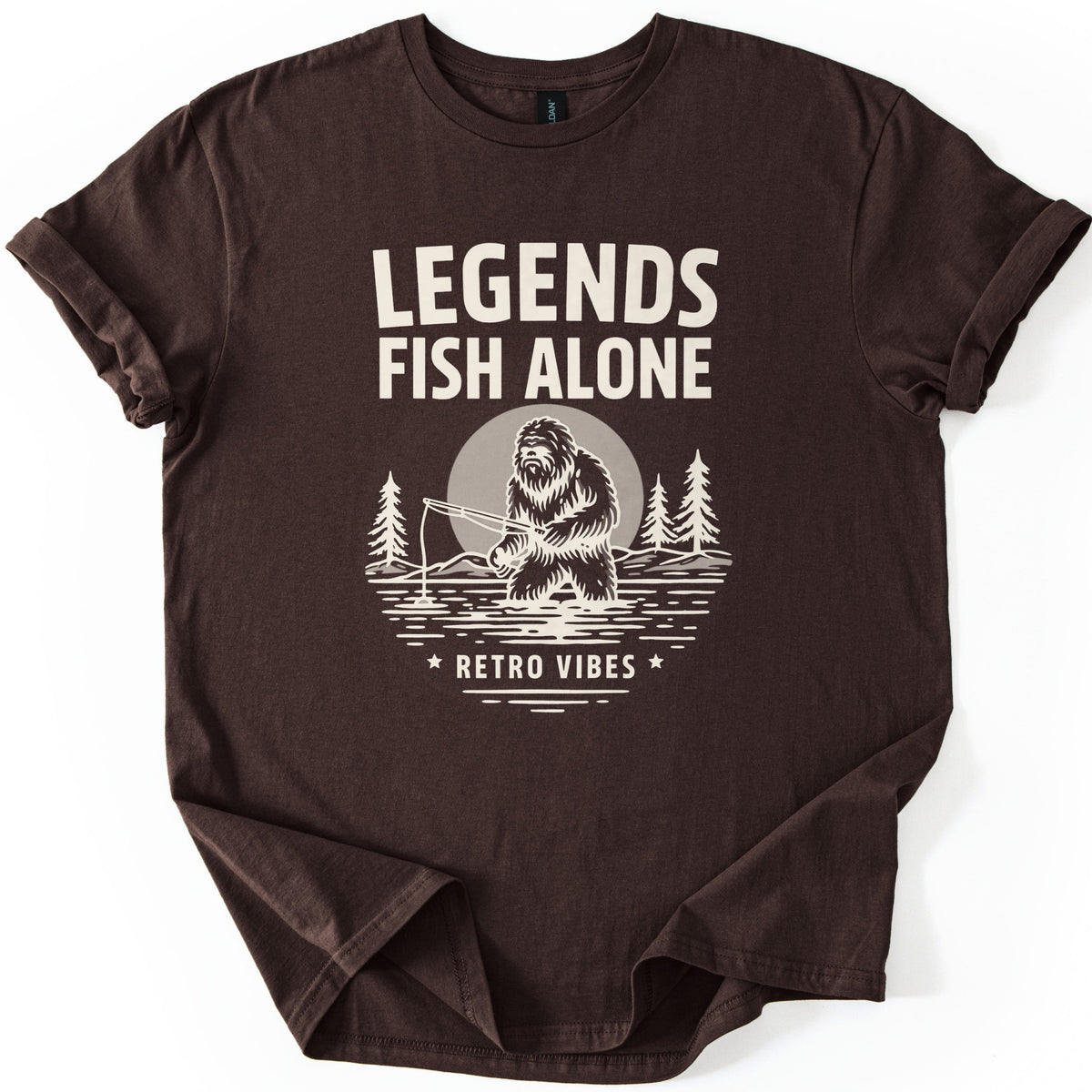 Legends Fish Alone Bigfoot Comfort Colors T-shirt in Dark Chocolate, laid flat. Cream print of Bigfoot fishing knee-deep in a lake before a gray moon and pine trees, under the words Legends Fish Alone and above Retro Vibes between two stars. Sleeves cuffed, on a white background.