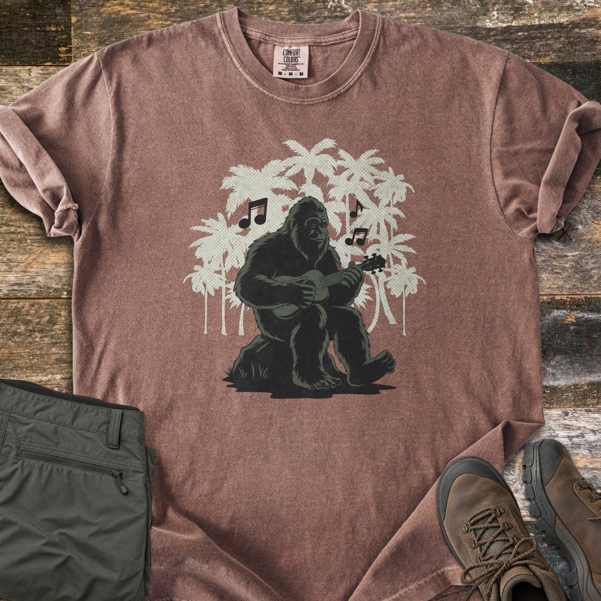 Bigfoot Ukulele Comfort Colors T-shirt in Espresso, laid flat. A Bigfoot sits on a rock playing a ukulele, with music notes and cream palm tree silhouettes behind him. Olive hiking pants and brown hiking boots lie beside it on weathered wood planks.