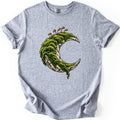 Moss Moon Comfort Colors T-shirt in Sport Grey, laid flat. A pale stone crescent moon covered in thick green moss, with thin orange moss sprouts rising from the top and moss trailing off the lower tip. No words. The sleeves are rolled on a white background.