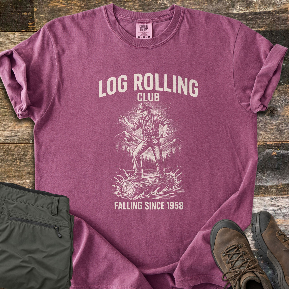 Log Rolling Club Comfort Colors T-shirt in Berry, laid flat. A sketched lumberjack in a hat and plaid shirt, holding an axe, balances on a log splashing through water below the words Log Rolling Club and above Falling Since 1958. Olive pants and brown boots sit on wood planks.
