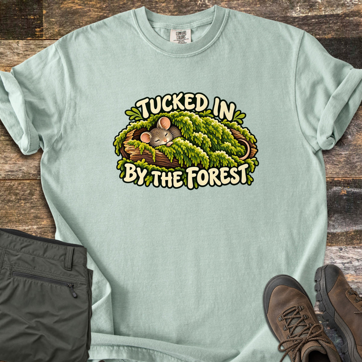 Tucked in by the Forest Comfort Colors T-shirt in Bay, laid flat. A sleeping cartoon mouse lies under a blanket of green moss in a hollow log between the words Tucked In By The Forest. Folded olive hiking pants and brown hiking boots sit on weathered wood planks.