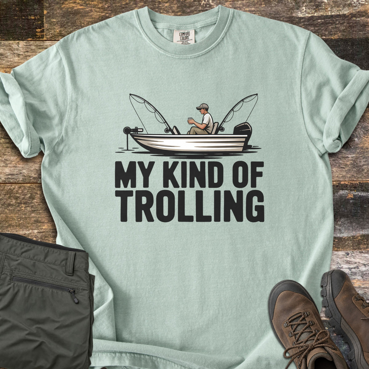 My Kind Of Trolling Comfort Colors T-shirt in Bay, laid flat. A man in a cap holds a cup in a small fishing boat with two bent rods and motors, above the words My Kind Of Trolling in black. Olive cargo pants and brown hiking boots sit beside it on weathered wood.