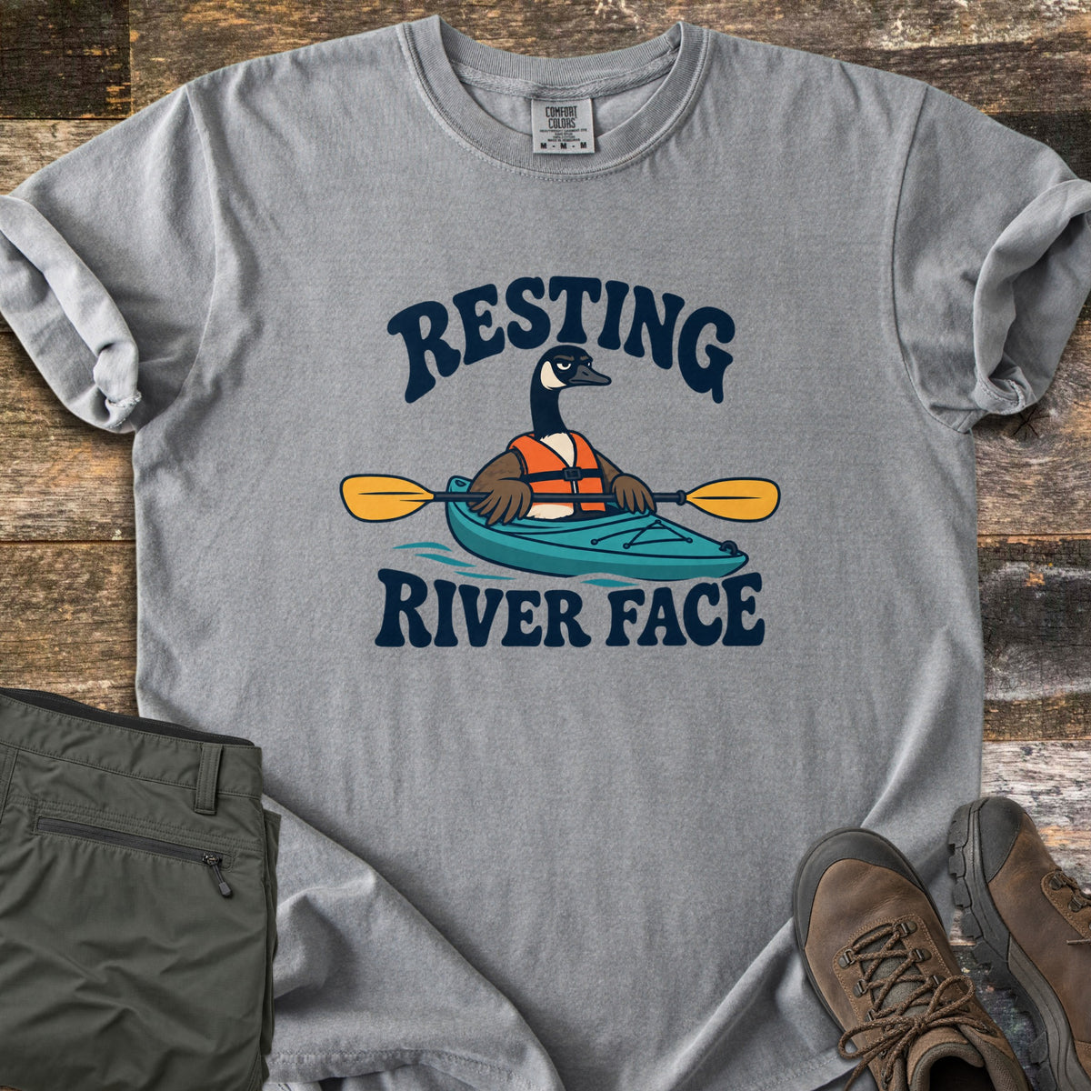 Resting River Face Comfort Colors T-shirt in Grey, laid flat. A grumpy cartoon Canada goose in an orange life vest paddles a teal kayak with a yellow paddle between the words Resting River Face. Olive hiking pants and brown hiking boots sit beside it on rustic wood.