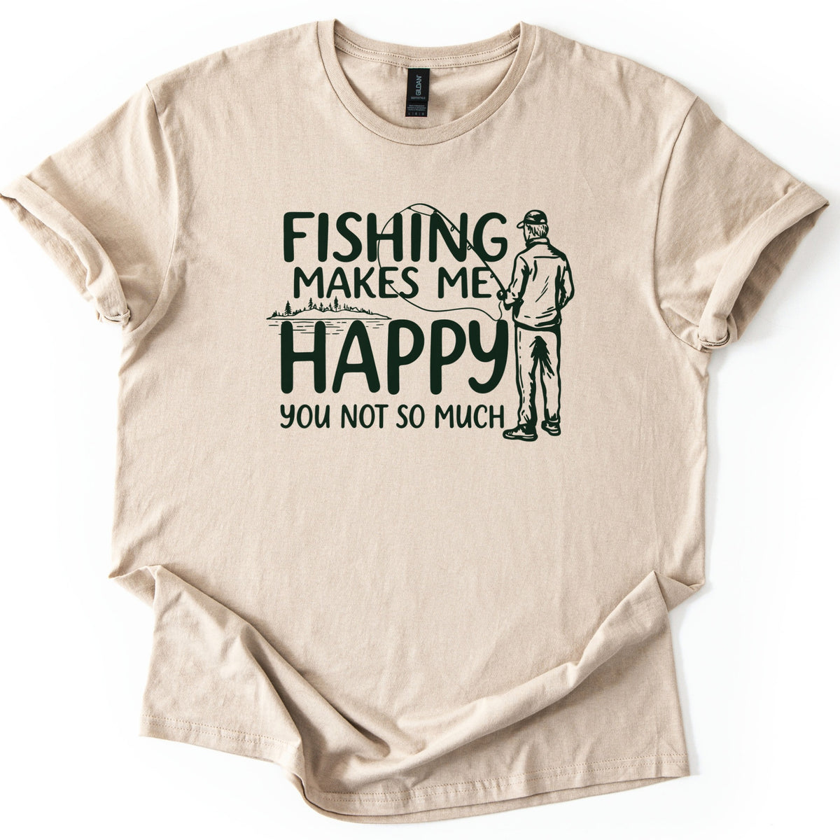 Fishing Makes Me Happy Comfort Colors T-shirt in Sand, laid flat. Dark green text reads Fishing Makes Me Happy, You Not So Much, beside a sketch of a man in a cap casting a rod from behind and a small lakeshore with pine trees. Sleeves are rolled on a white background.