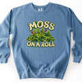 Moss on a Roll Comfort Colors crewneck sweatshirt in Blue Jean, laid flat. Cream bubble letters reading MOSS ON A ROLL frame a mound of green moss and fern fronds made of rolled paper-quilling swirls, with two rust-brown spore capsules. It lies on a plain white surface.