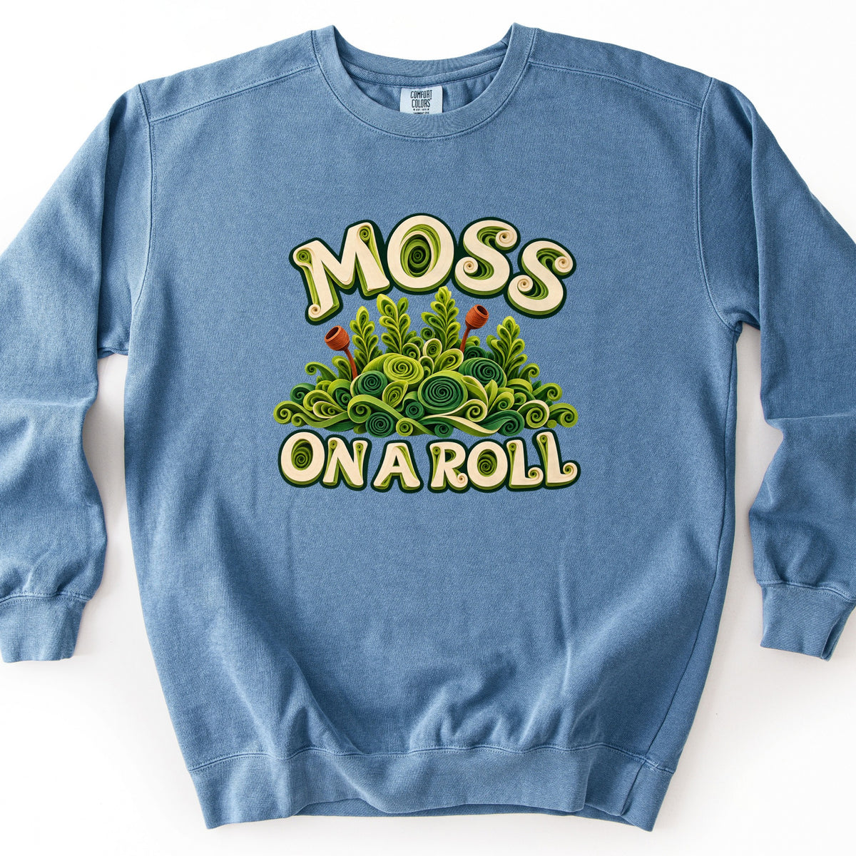 Moss on a Roll Comfort Colors crewneck sweatshirt in Blue Jean, laid flat. Cream bubble letters reading MOSS ON A ROLL frame a mound of green moss and fern fronds made of rolled paper-quilling swirls, with two rust-brown spore capsules. It lies on a plain white surface.