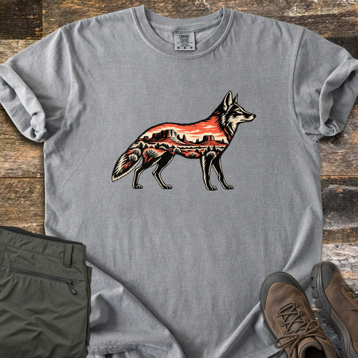 Desert Mesa Coyote Comfort Colors T-shirt in Grey, laid flat. A side-view coyote silhouette filled with a red desert mesa landscape, sunset sky and shrubs, with no words. Sleeves rolled on rustic wood, beside folded olive hiking pants and brown hiking boots.