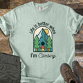 Better When I'm Climbing Comfort Colors T-shirt in Bay, laid flat. An arched scene of a person facing a mountain with a winding trail, pine trees and a yellow sun, with the words Life is better when I'm Climbing. Gray pants and brown hiking boots lie on a wood floor.