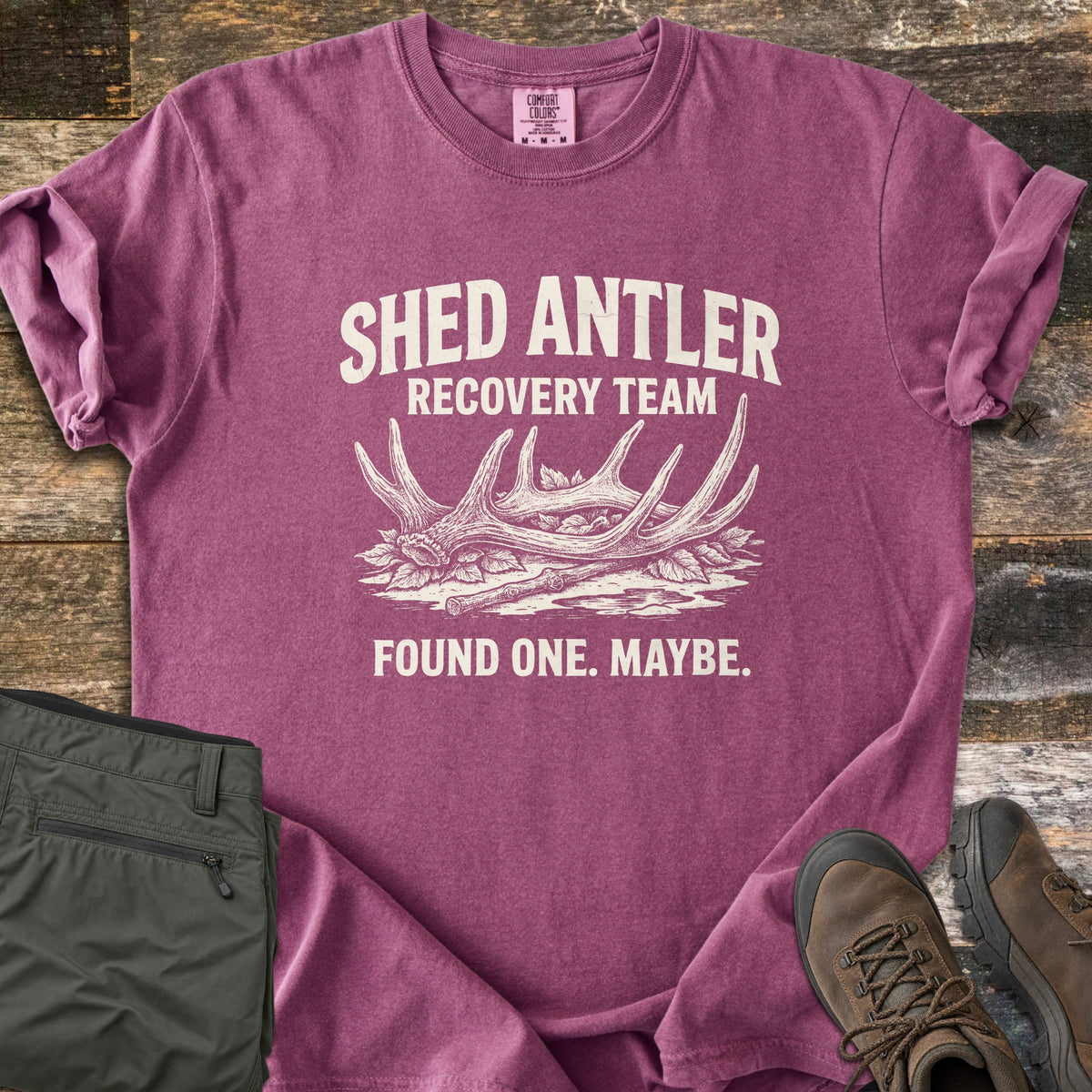 Shed Antler Recovery Team Comfort Colors T-shirt in Berry, laid flat. A sketched shed antler rests on leaves beside a stick under the words Shed Antler Recovery Team, with Found One. Maybe. below. Olive pants and brown hiking boots sit beside it on weathered wood.