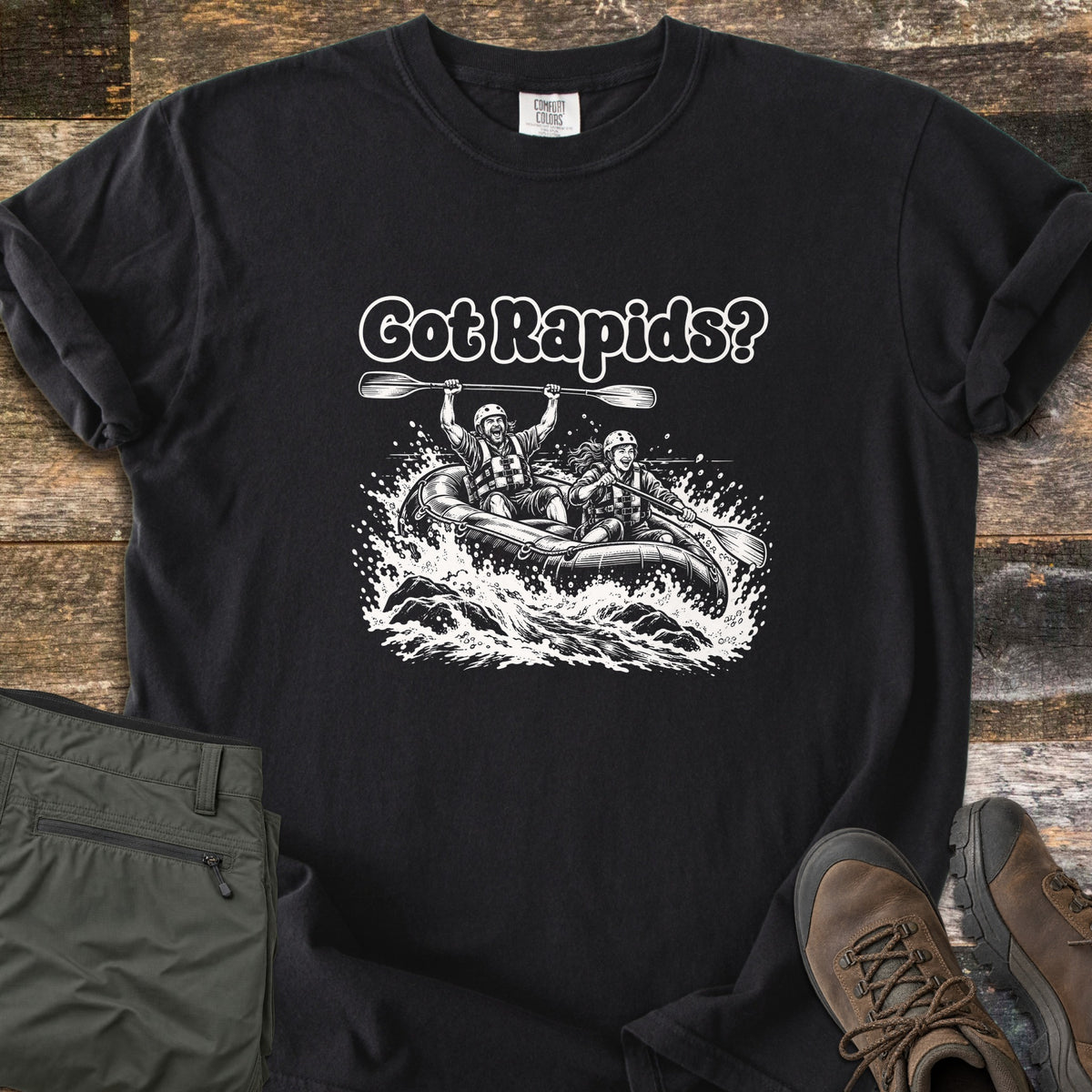 Got Rapids Comfort Colors T-shirt in Black, laid flat. The words Got Rapids? above two smiling rafters in helmets and life vests riding a raft through splashing whitewater, one holding a paddle overhead. Set on wood planks with olive hiking pants and brown boots.