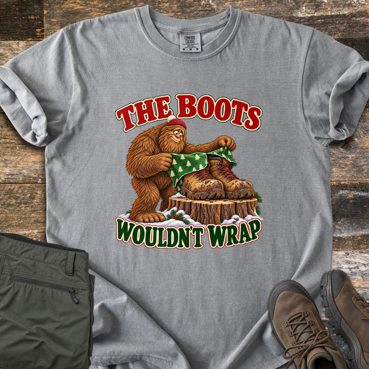 The Boots Wouldn't Wrap Comfort Colors T-shirt in Grey, laid flat. A Bigfoot in a red beanie tries to wrap muddy hiking boots on a snowy stump in green Christmas tree paper, between the words The Boots Wouldn't Wrap. Olive pants and brown hiking boots lie beside it on weathered wood.