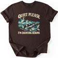 Counting Herons Comfort Colors T-shirt in Dark Chocolate, laid flat. Three herons wade in marsh grass beside a winding river with an empty kayak and paddle, drawn in teal and cream, between the words Quiet Please and I'm Counting Herons. Sleeves are rolled on a white background.