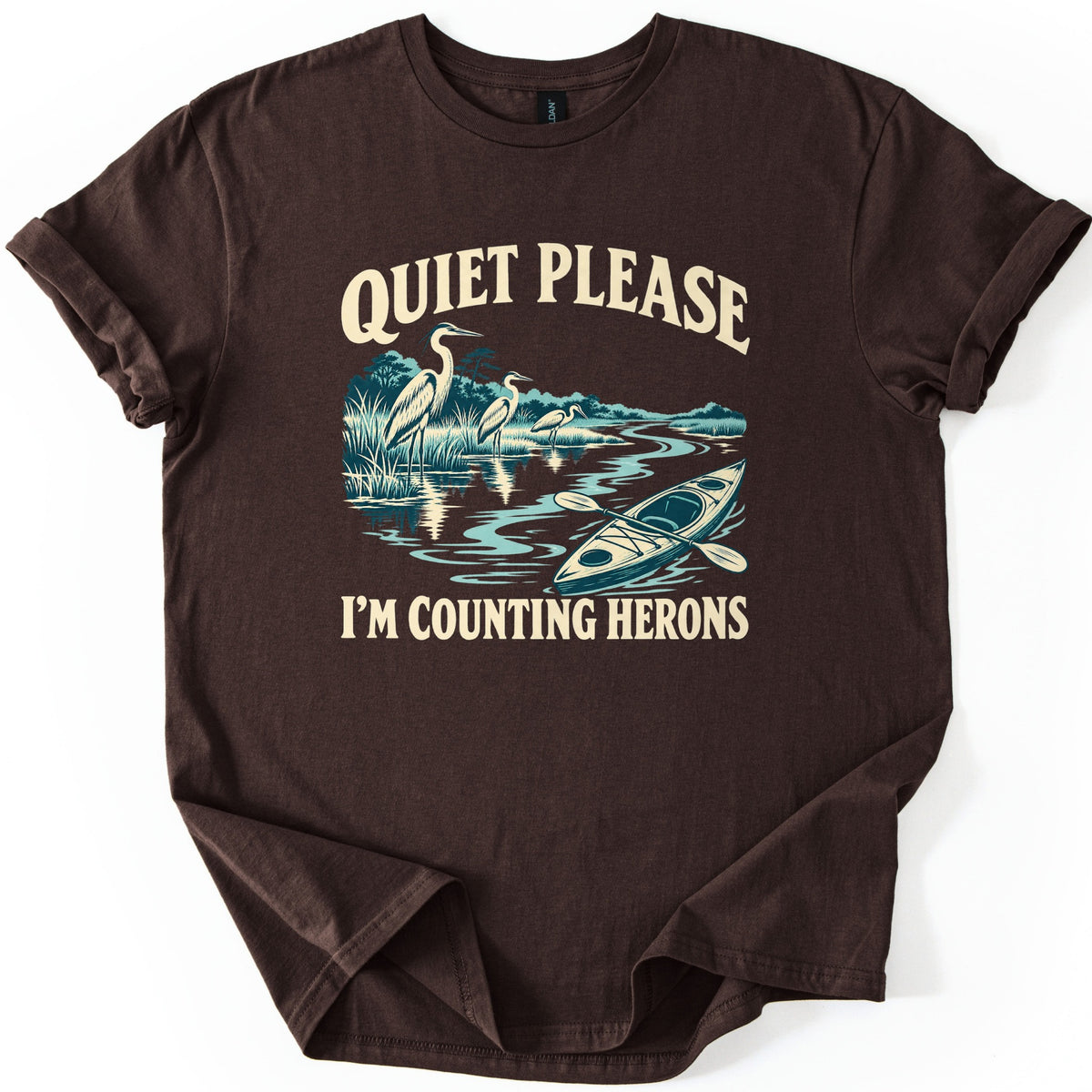 Counting Herons Comfort Colors T-shirt in Dark Chocolate, laid flat. Three herons wade in marsh grass beside a winding river with an empty kayak and paddle, drawn in teal and cream, between the words Quiet Please and I'm Counting Herons. Sleeves are rolled on a white background.