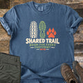 Shared Trail Room for Every Footprint Comfort Colors T-shirt in Denim, laid flat. A bike tire track, a boot print and a paw print sit above the words Shared Trail, Room for Every Footprint, MTB. Folded olive hiking pants and brown hiking boots lie beside it on dark weathered wood.