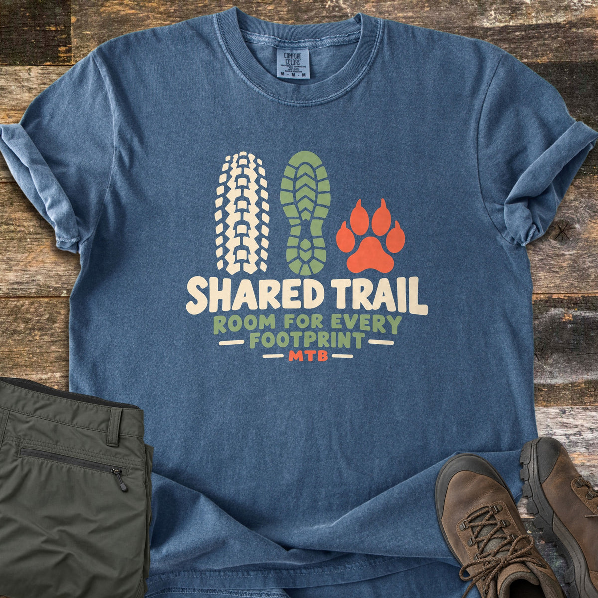Shared Trail Room for Every Footprint Comfort Colors T-shirt in Denim, laid flat. A bike tire track, a boot print and a paw print sit above the words Shared Trail, Room for Every Footprint, MTB. Folded olive hiking pants and brown hiking boots lie beside it on dark weathered wood.