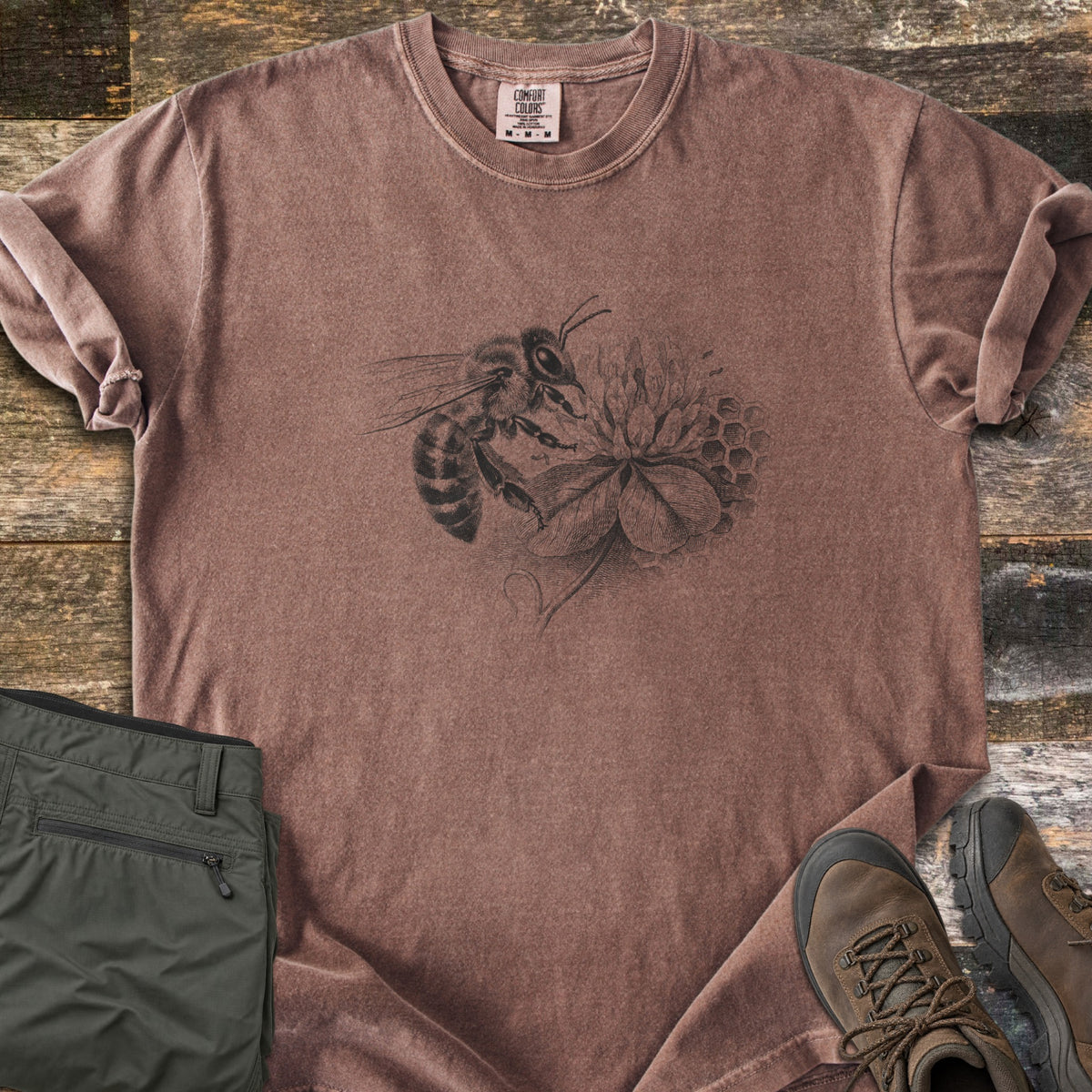 Honeybee On Clover Comfort Colors T-shirt in Espresso, laid flat. A detailed black engraving-style illustration of a honeybee on a clover blossom with honeycomb cells behind it, no words. Sleeves rolled, on weathered wood planks beside olive hiking pants and brown hiking boots.