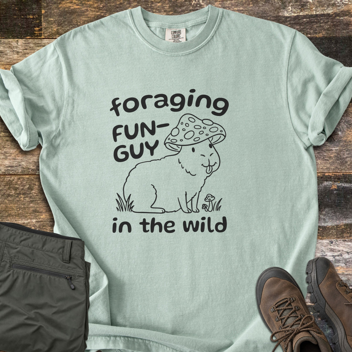 Foraging Fun-Guy in the Wild Comfort Colors T-shirt in Bay, laid flat. A black line drawing of a capybara wearing a spotted mushroom cap, tongue out, sits in grass by tiny mushrooms between the words foraging FUN-GUY in the wild. Olive hiking pants and brown boots lie on weathered wood.