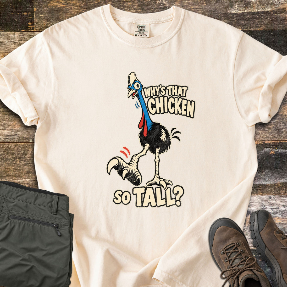 Why's That Chicken So Tall Comfort Colors T-shirt in Ivory, laid flat. A cartoon cassowary with a blue neck, red wattles and a huge clawed foot raised mid-step, with the words Why's That Chicken So Tall? Folded green cargo pants and brown hiking boots sit on weathered wood.