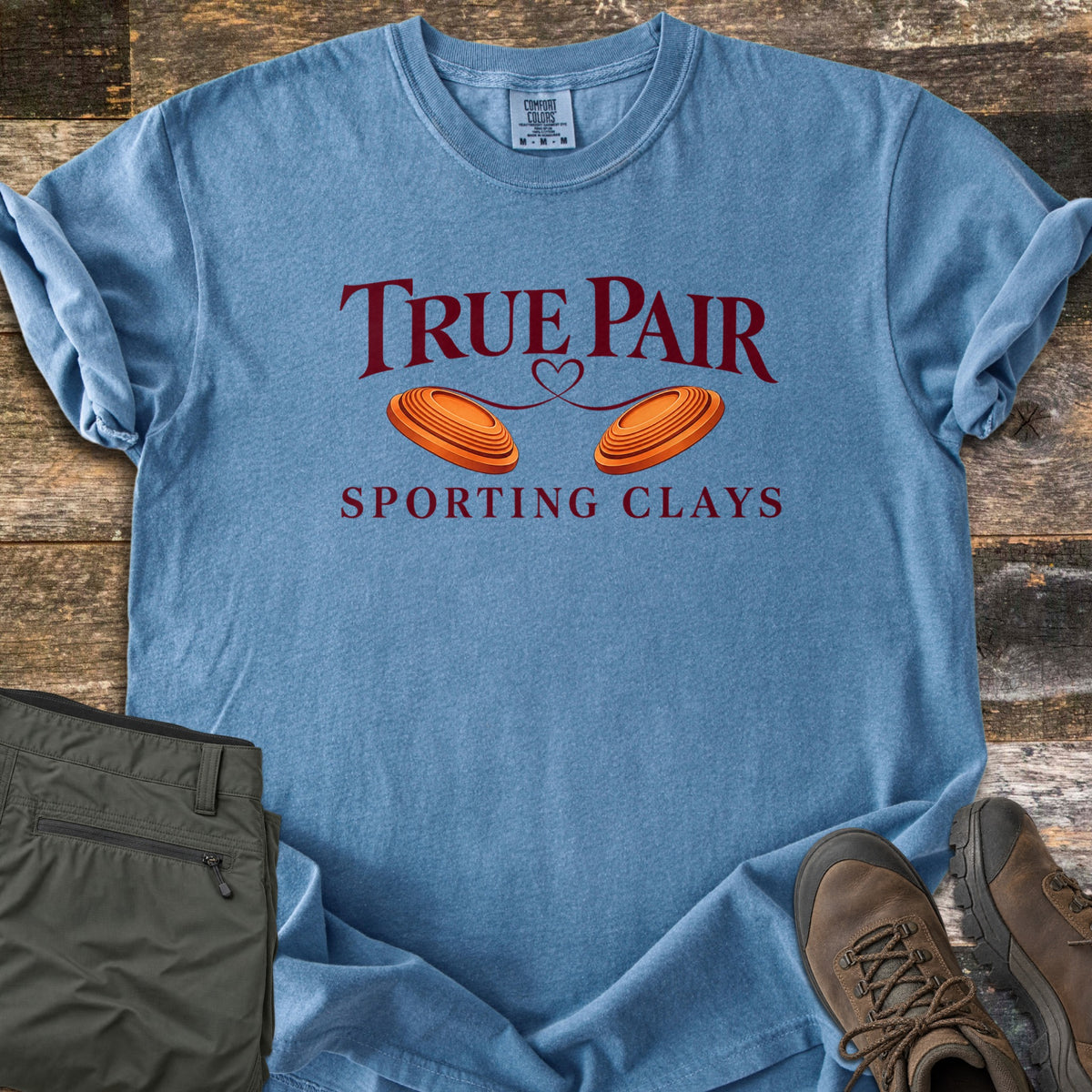True Pair Comfort Colors T-shirt in Blue Jean, laid flat. Dark red text reads True Pair above two orange clay targets joined by a looping heart line, with Sporting Clays below. Sleeves are rolled; olive pants and brown hiking boots sit on weathered wood.