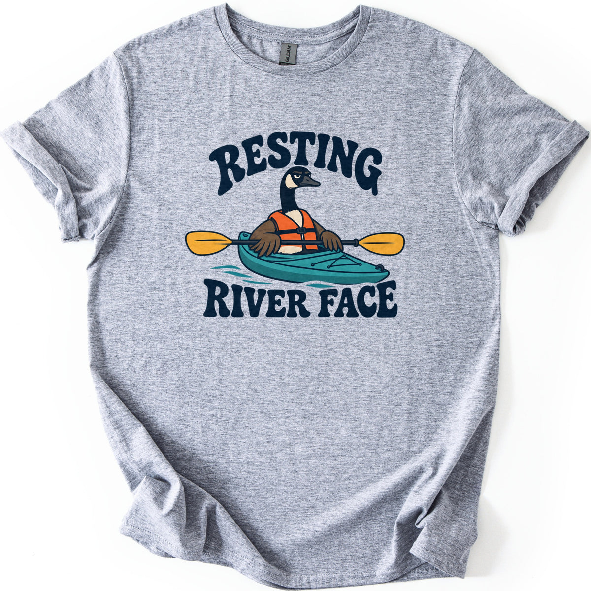 Resting River Face Comfort Colors T-shirt in Sport Grey, laid flat. A scowling cartoon Canada goose in an orange life vest paddles a teal kayak with a yellow paddle, between the words RESTING above and RIVER FACE below. The shirt lies on a white background with cuffed sleeves.