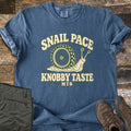 Snail Pace Knobby Taste Comfort Colors T-shirt in Denim, laid flat. A cartoon snail whose shell is a knobby mountain bike tire sits between the words Snail Pace and Knobby Taste, with MTB below. It lies on weathered wood beside folded olive hiking pants and brown hiking boots.
