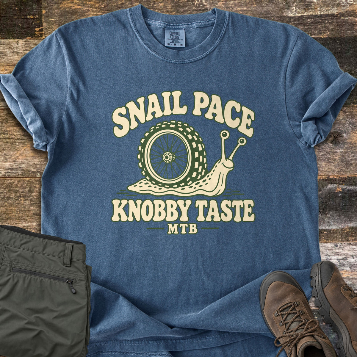 Snail Pace Knobby Taste Comfort Colors T-shirt in Denim, laid flat. A cartoon snail whose shell is a knobby mountain bike tire sits between the words Snail Pace and Knobby Taste, with MTB below. It lies on weathered wood beside folded olive hiking pants and brown hiking boots.